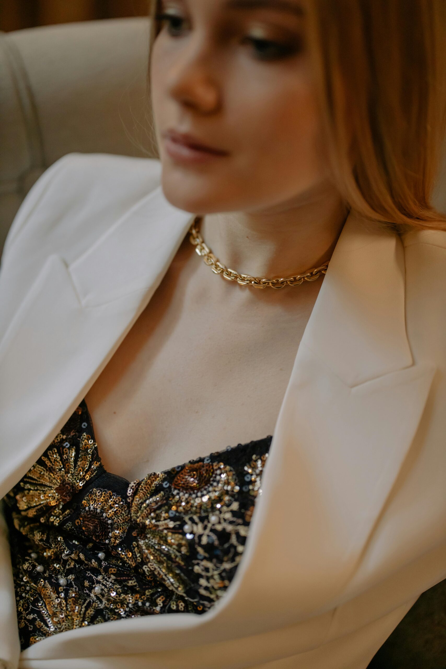 A stylish woman wearing a gold necklace and embellished top with a white blazer indoors.